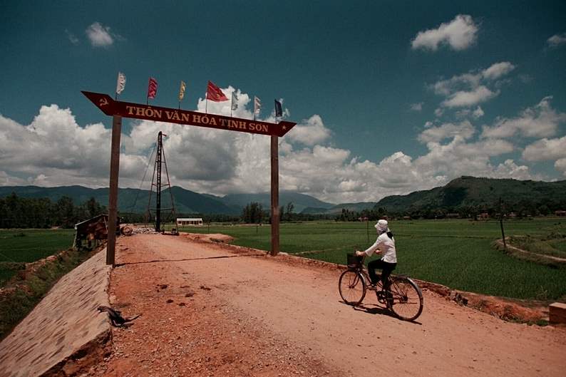 A-woman-pedals-her-bicycle-down-a-rural-road-near-Chu-Lai