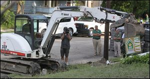 LeBron James operates an excavator as a camera operator and Darren Rowland and Brian Rice of Rice Nursery and Landscaping, look on. 